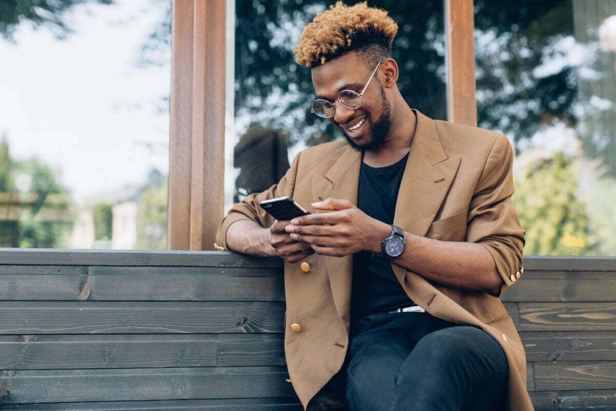 A young African American man smiles while using his phone on a park bench