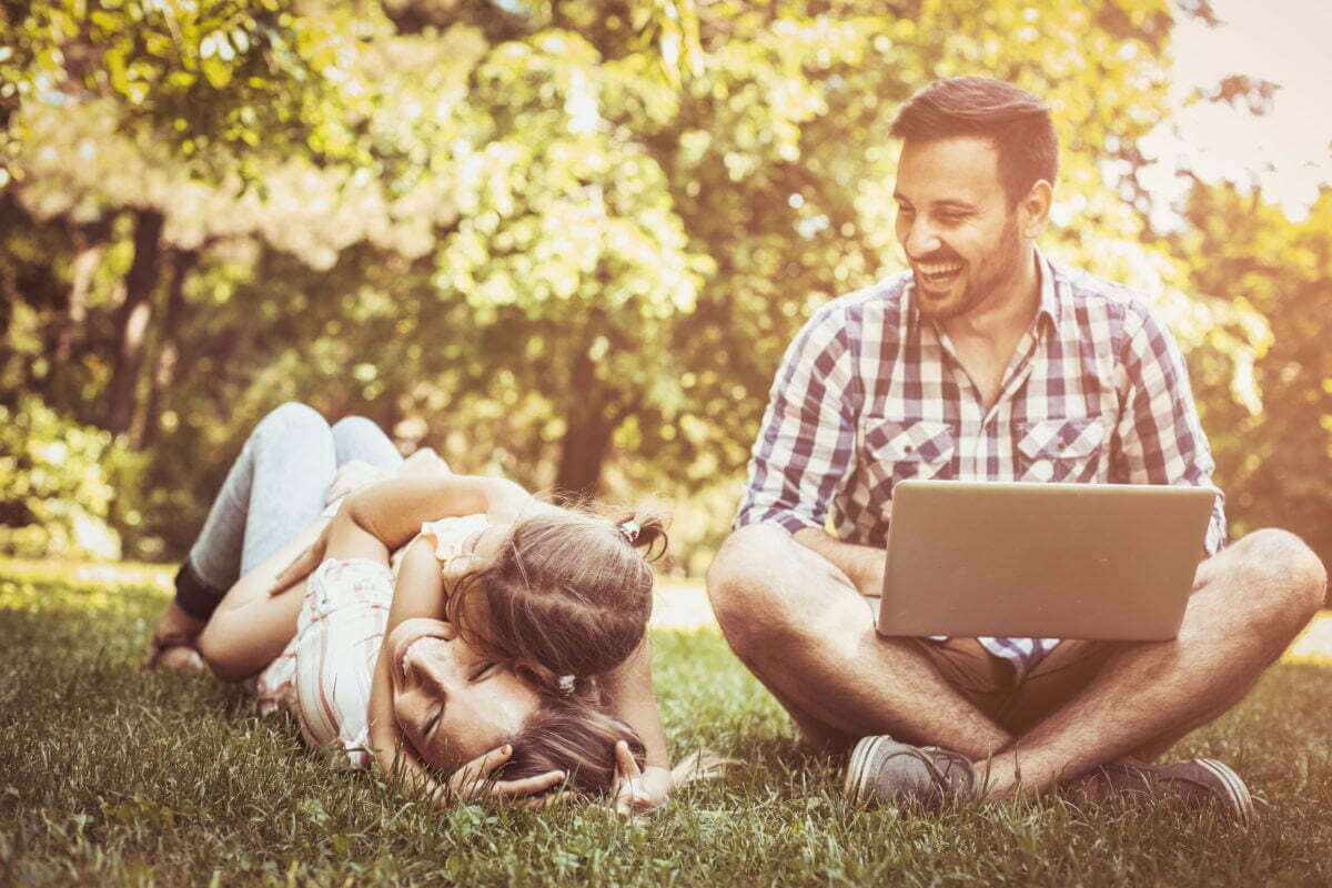 Father with laptop smiles as his family plays in park